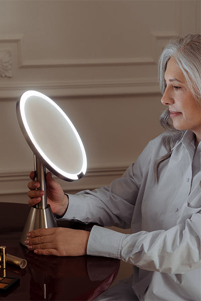 Woman using a handheld premium makeup mirror with LED lights in an elegant room.
