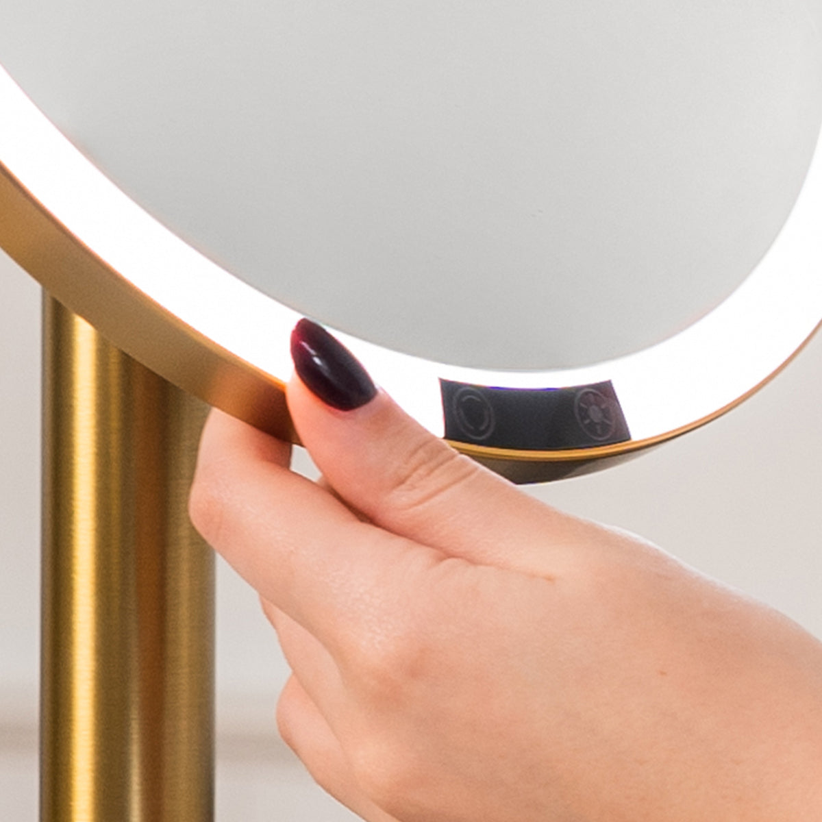 Hand adjusting a round lighted 5x magnification mirror with a gold frame.