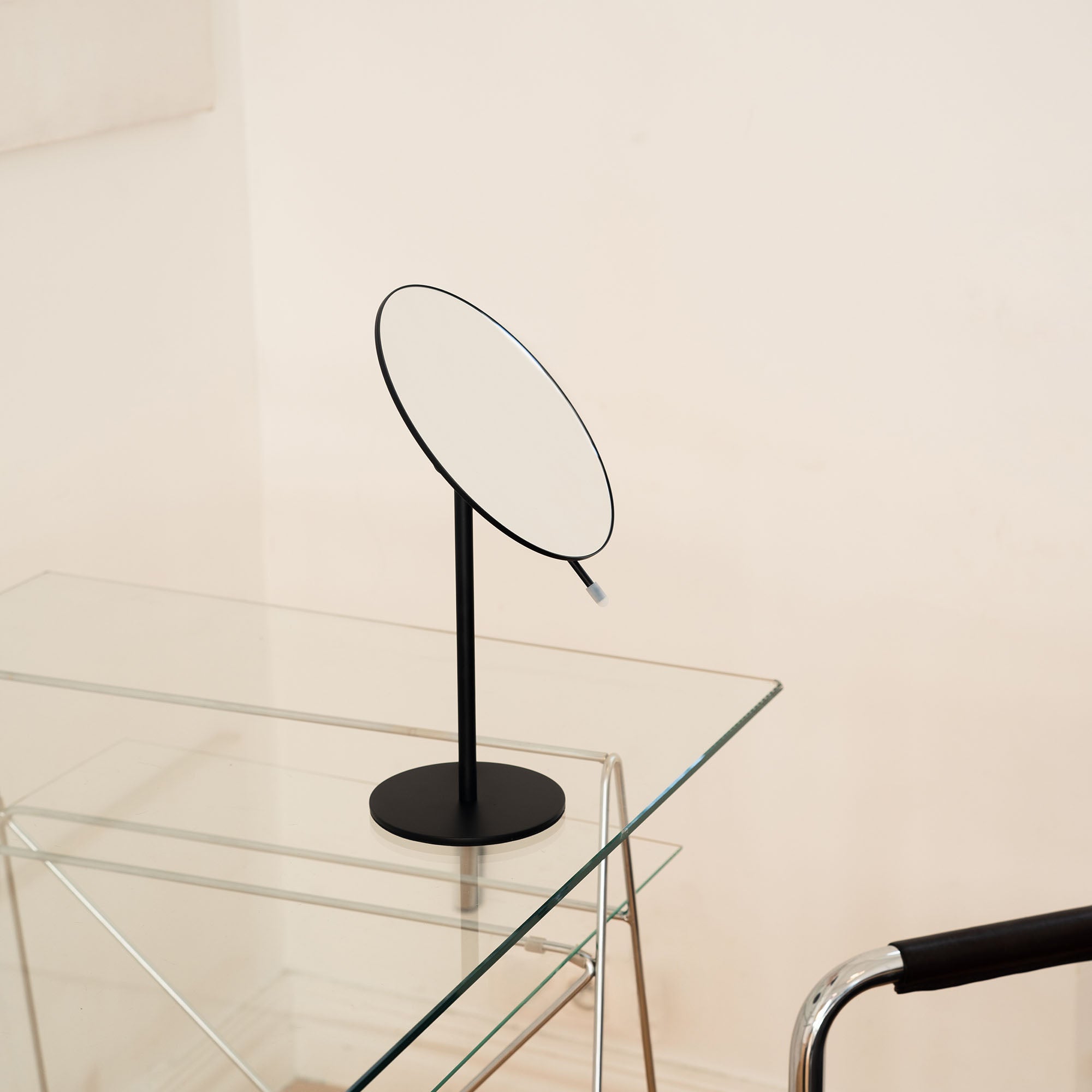 Black round mirror on a stand next to a glass table with a chair in a minimalistic setting_#Silver_Black