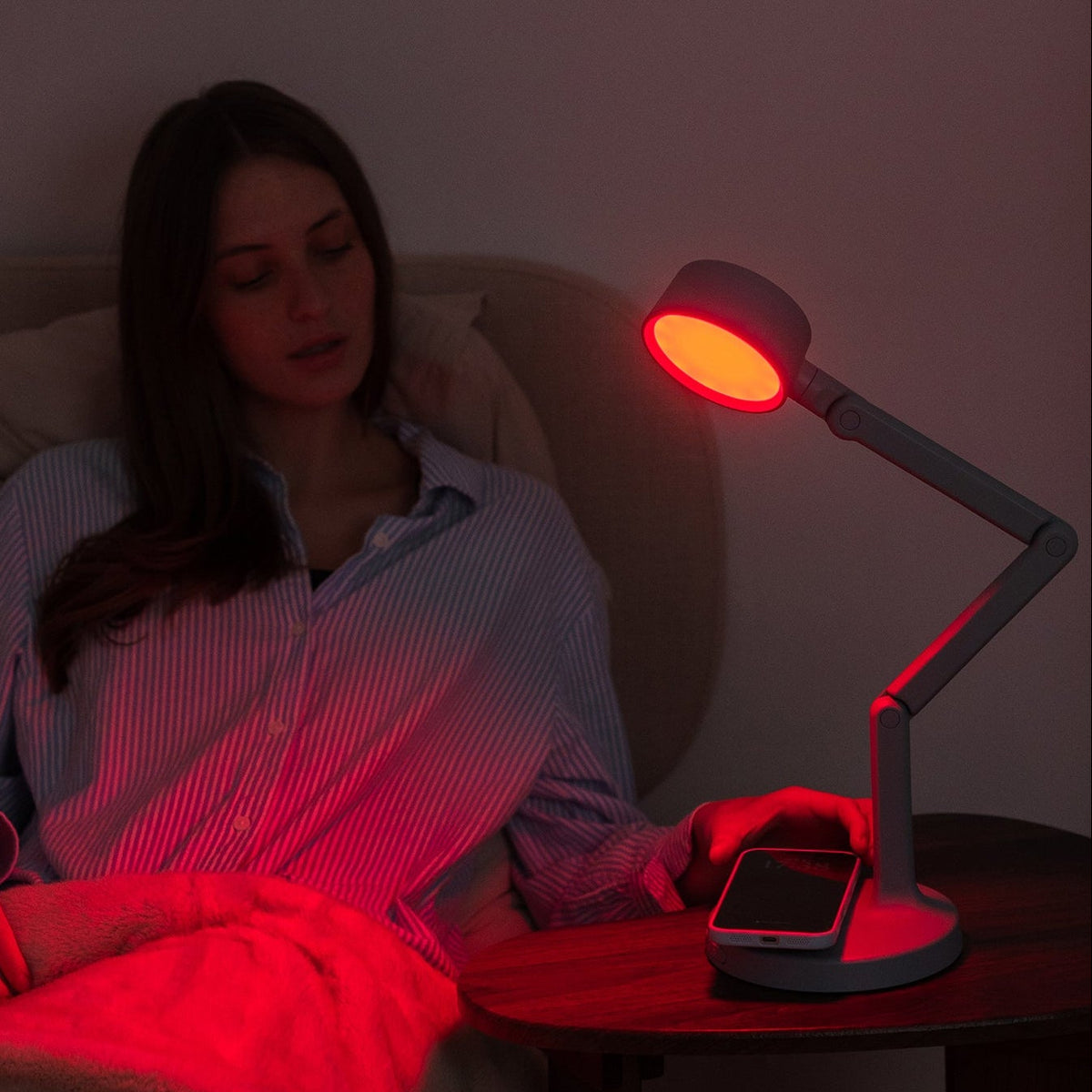 A woman in a striped pajama shirt sits in bed under a blanket, using a bedside table lamp with a red light therapy head to read her phone on a dark wooden nightstand.