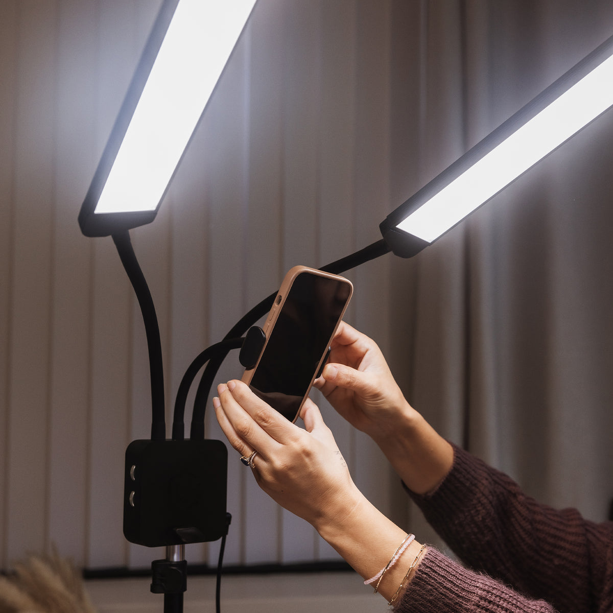 A person holding a smartphone in front of two bright LED light panels mounted on a stand, with a softly lit indoor background.