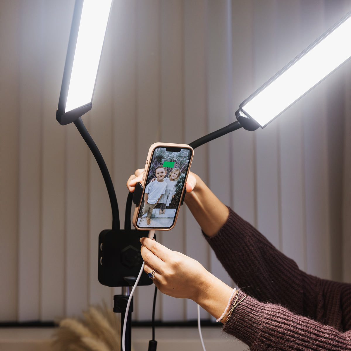 A person holds a smartphone between two bright LED light panels mounted on flexible black arms. The phone is connected to a black wireless charging stand, with a phone photo visible on the screen.