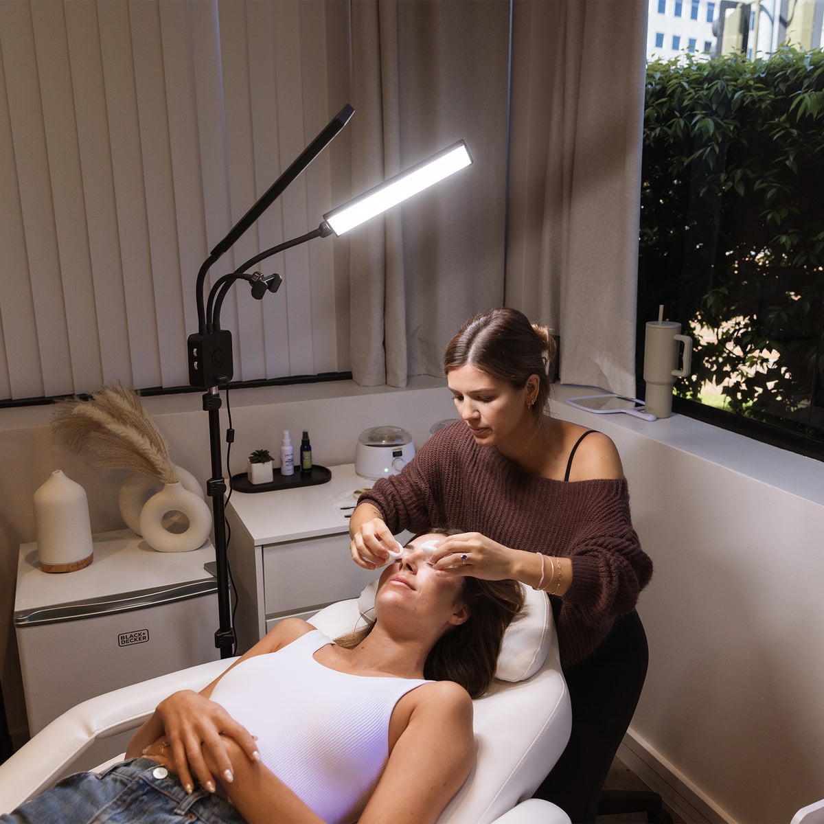 A woman receives a facial treatment while reclining in a spa chair, with a practitioner standing beside her under a bright adjustable LED lamp in a modern treatment room.