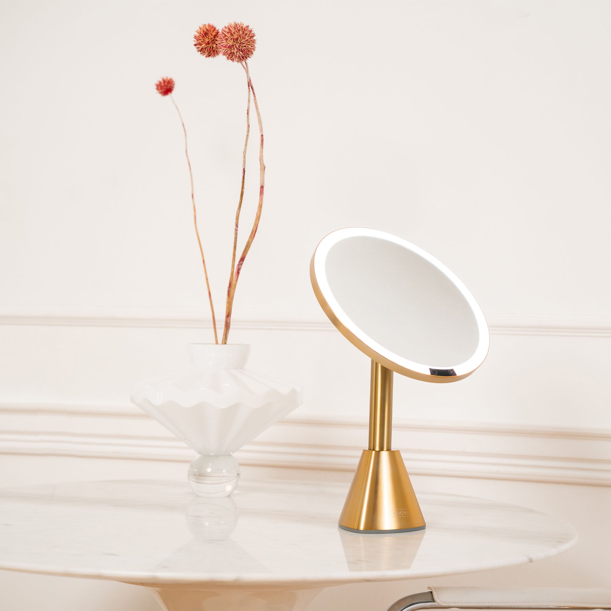 Gold vanity mirror on a light wooden surface with a vase of flowers in the background.