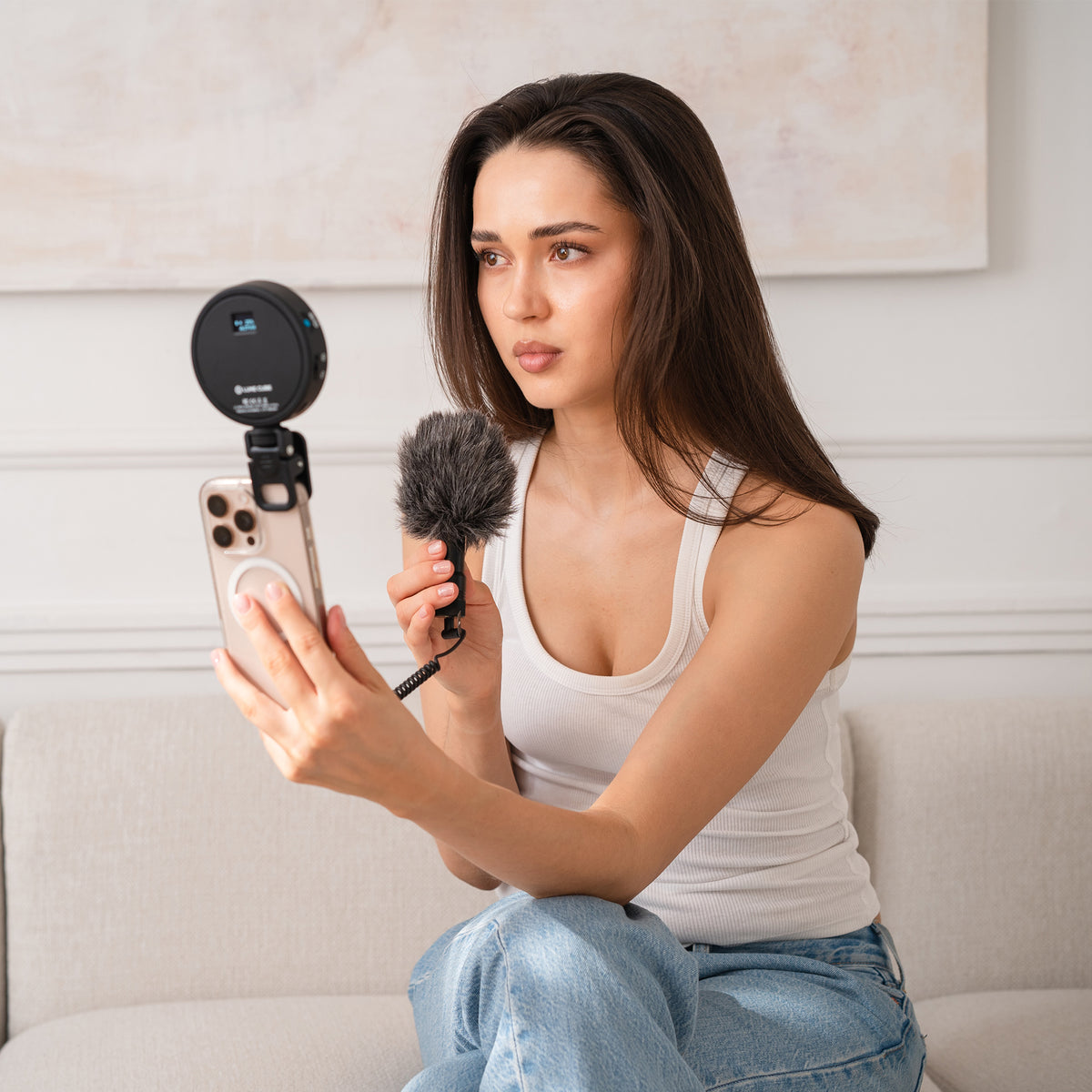 A woman sitting on a light beige sofa holds a smartphone with a phone mount and circular microphone attachment, while also holding a fuzzy handheld microphone. She is wearing a white tank top and blue jeans in a bright, modern room with a framed painting on the wall.