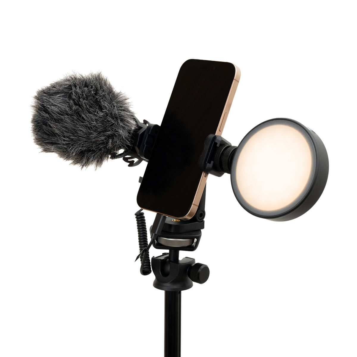 A studio setup with a smartphone in a vertical phone mount, a gray furry microphone windscreen on the left, and a round ring light on the right, all on a black stand against a plain white background.