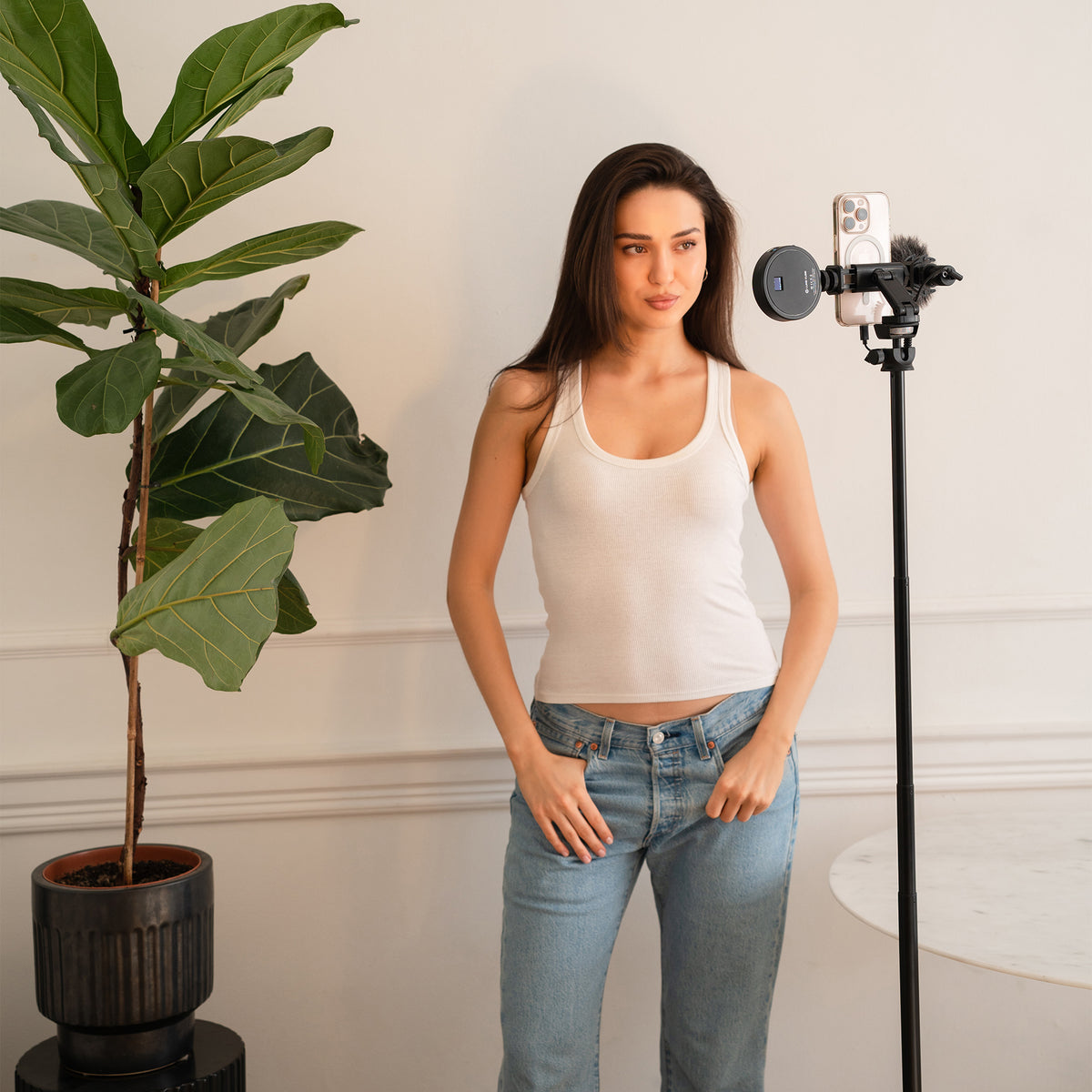 A woman wearing a white ribbed tank top and light blue jeans stands indoors next to a phone mounted on a stand, with a large potted fiddle-leaf fig plant on the left and a cream-colored wall behind her.