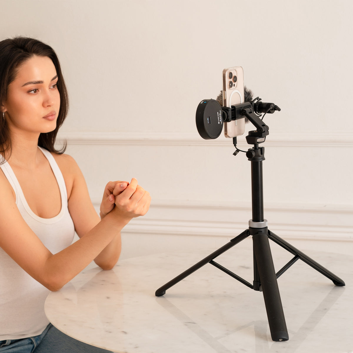A woman in a white tank top sits at a white marble table, looking at a smartphone mounted on a black tripod with a circular black accessory attached. The setting is a clean, minimalist room with white walls and molding.