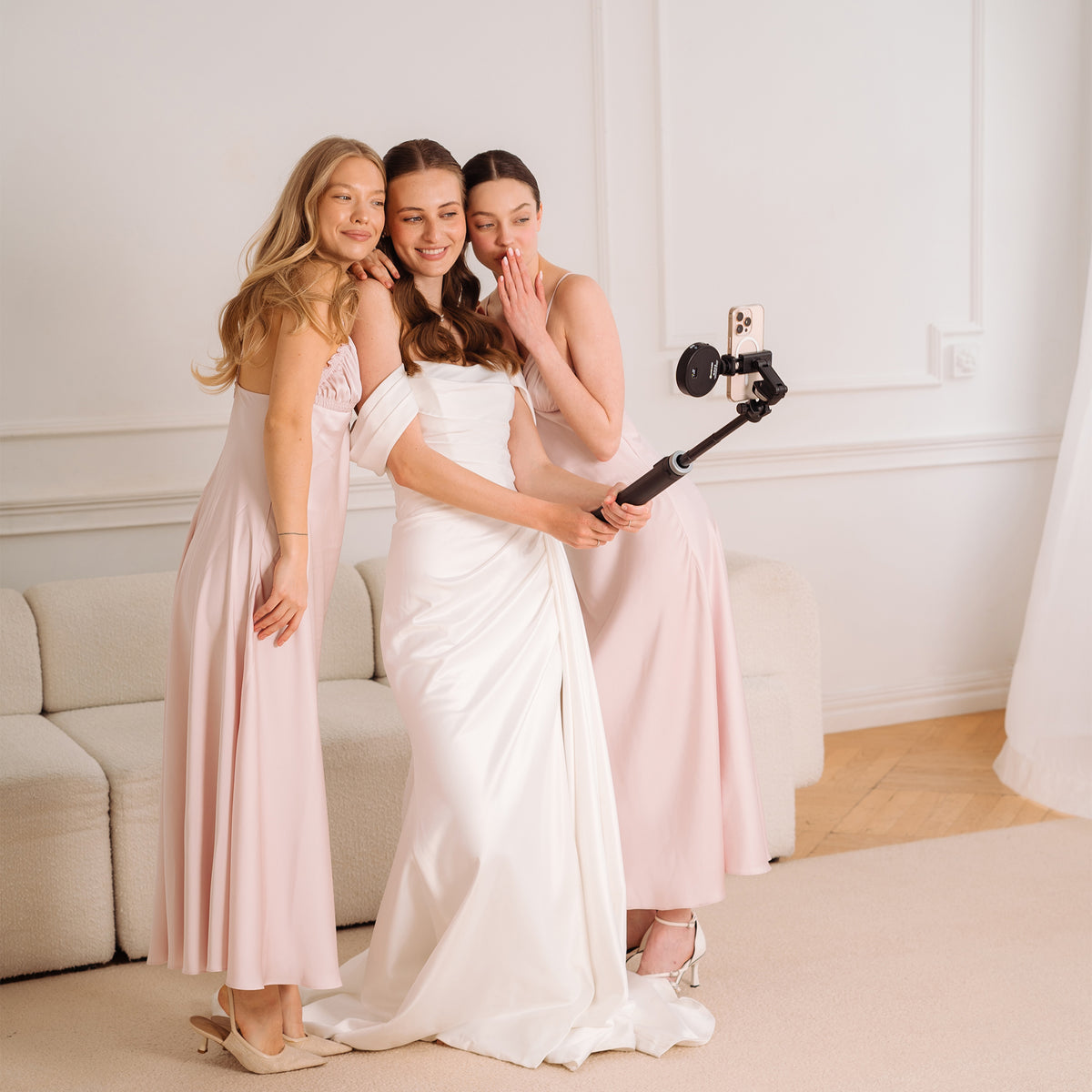 Three women posing together indoors, taking a selfie with a phone on a selfie stick. The woman in the center wears a white satin bridal gown, while the two women on either side wear matching pale blush pink bridesmaid dresses.