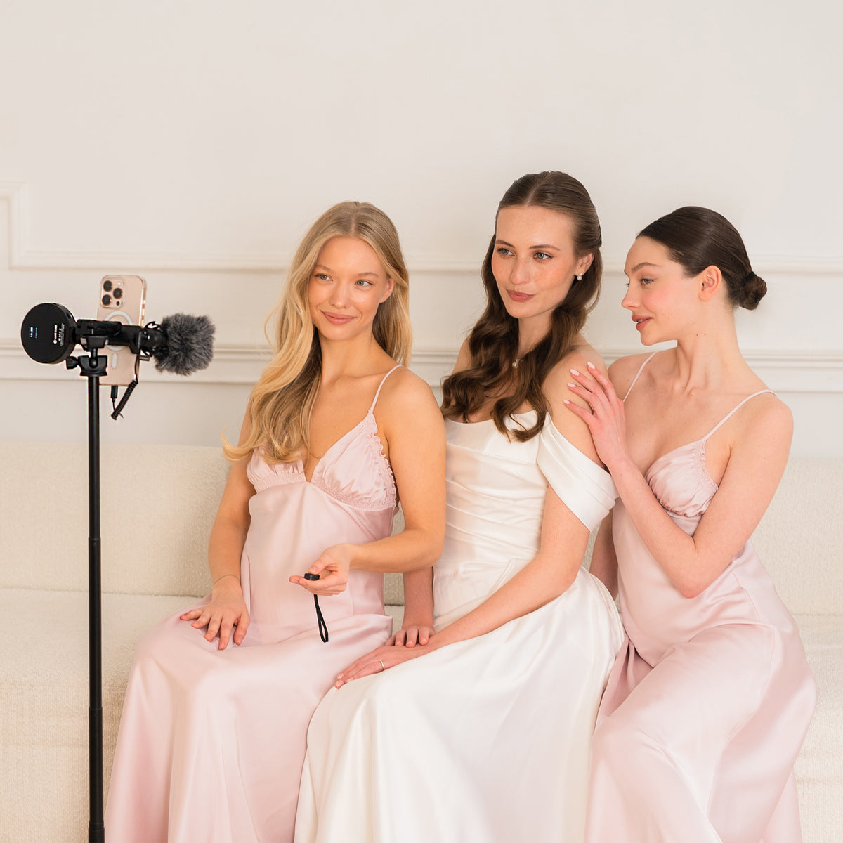 Three women in elegant satin bridesmaid gowns sit together on a white sofa, with a phone and microphone setup visible on the left. The dresses are in soft blush pink, white, and pale pink tones against a clean white interior background.
