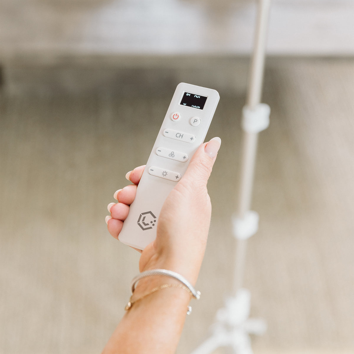 A hand holding a white remote control with a small digital display and control buttons, photographed against a softly blurred indoor background.