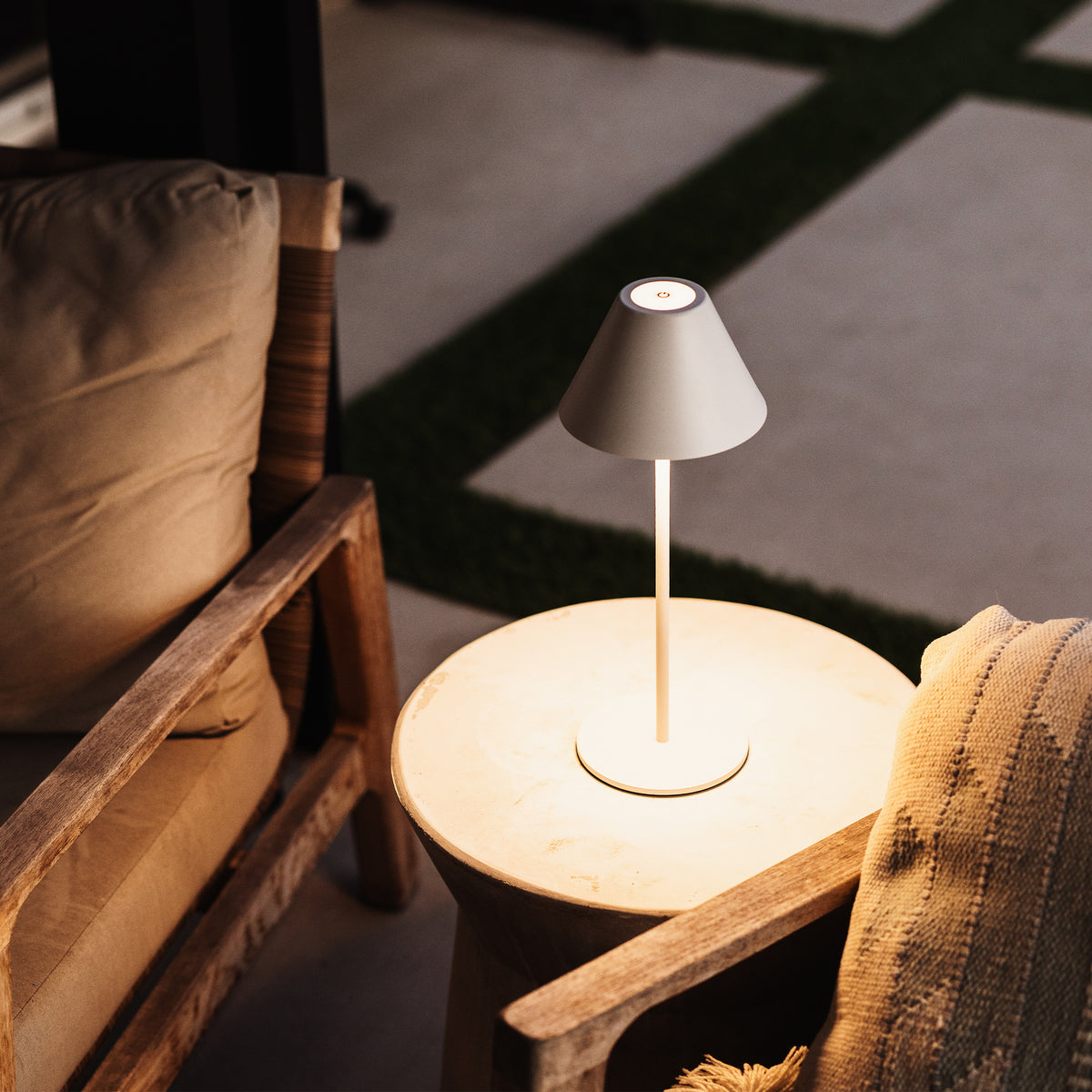 A modern table lamp with a tapered gray shade and a slim white stem, glowing on a round stone side table between two wooden armchairs in a warm indoor setting.