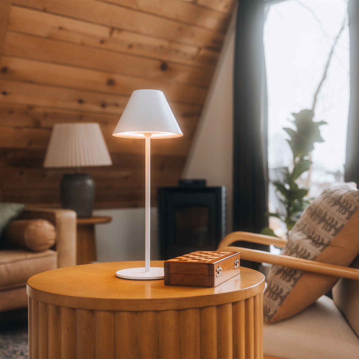 A modern white table lamp with a tapered shade and slim stem stands on a round wooden side table in a cozy living room. A small wooden chess box sits beside it, with a sofa, patterned cushion, fireplace, and window in the background.