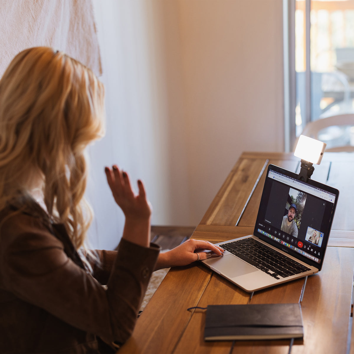 A woman with blonde hair sits at a wooden desk, seen from behind, using a laptop on a video call in a warm, softly lit room.