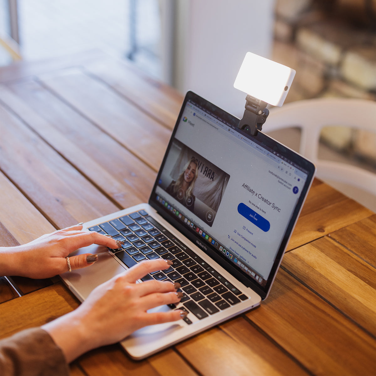 A person uses a silver laptop on a wooden table while wearing a white rectangular clip-on light attached to the laptop's screen, in a bright indoor setting.