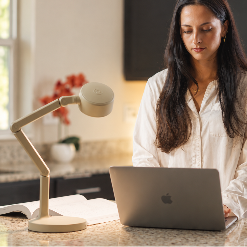 A woman in a white button-up shirt works at a kitchen island with an open laptop, a beige adjustable desk lamp, and a white mug on a speckled granite countertop.