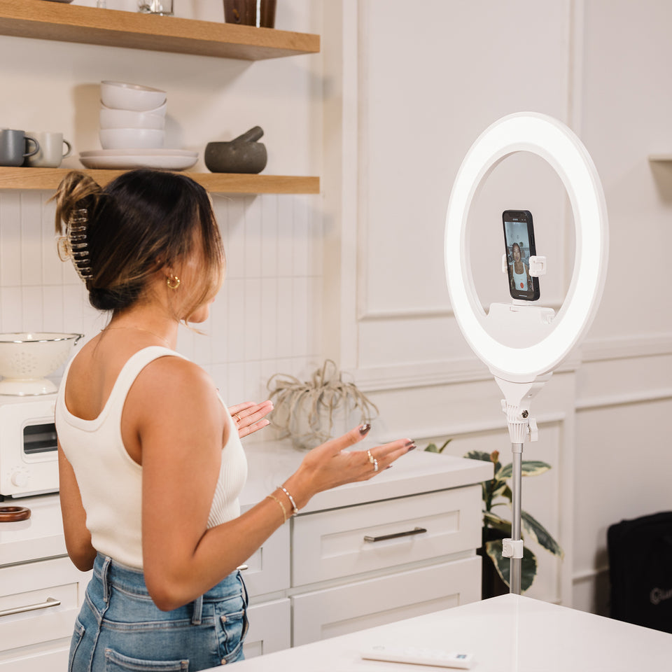 A woman stands in a bright kitchen facing a white ring light on a stand with a smartphone mounted inside the ring, appearing to record or stream.