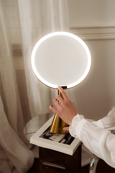 Person holding a round illuminated cordless lighted makeup mirror with a gold base.