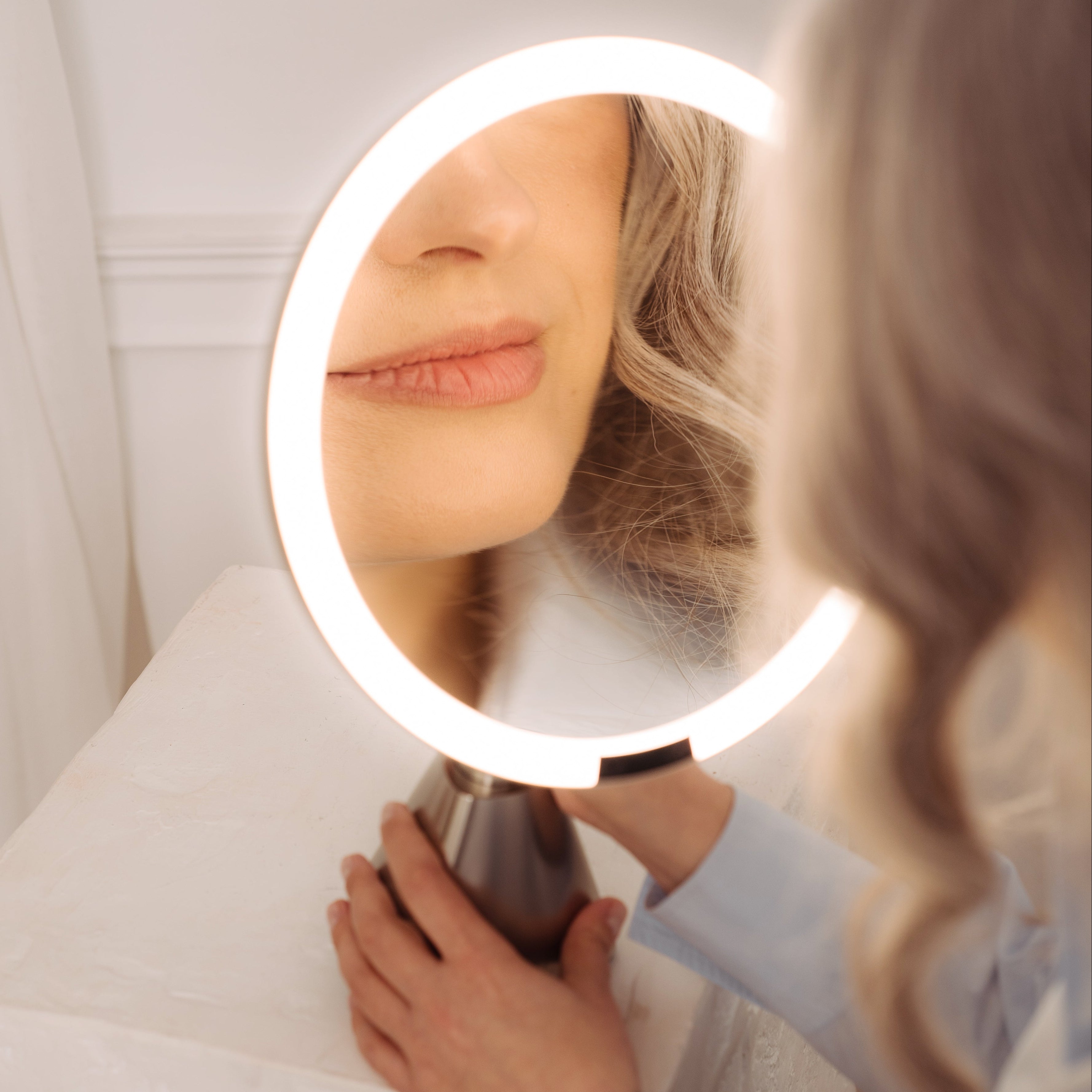 A woman with blonde hair looks into a circular LED vanity mirror with a glowing white ring, resting on a white pedestal in a bright indoor setting.