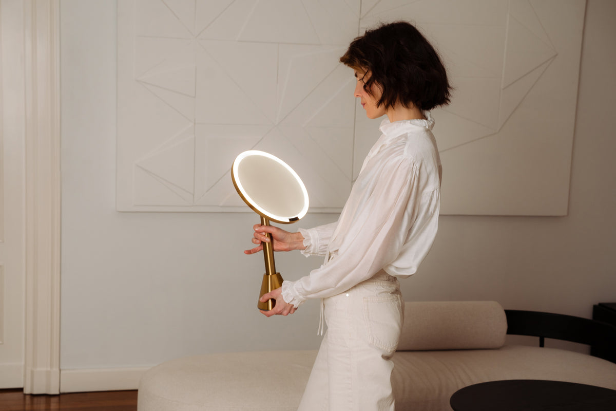 Woman holding a handheld mirror in a modern room with a bed and wall art.