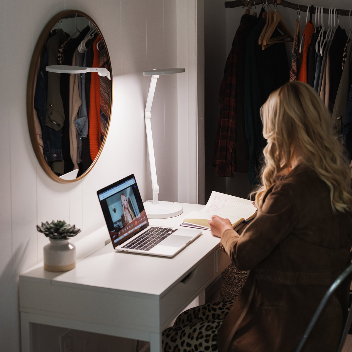 A woman sits at a white desk in a home office setting, illuminated by a modern white desk lamp. On the desk is an open laptop, papers, and a small potted succulent.