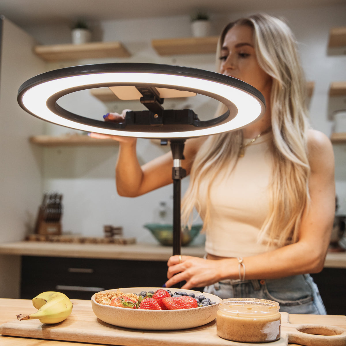 A woman in a kitchen setting holds a black ring light above a wooden cutting board with a bowl of fruit and a jar of peanut butter.