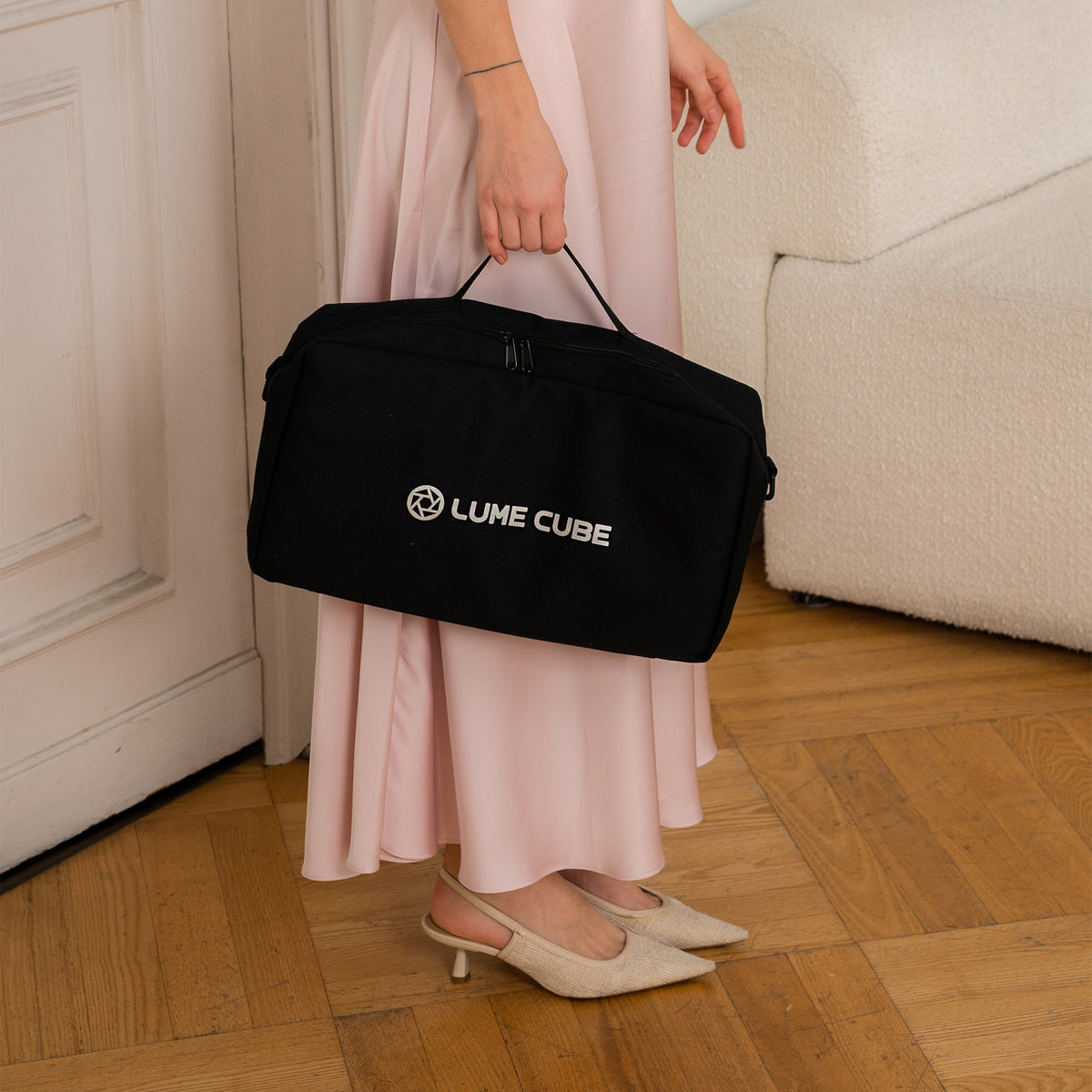 A person in a long light pink dress holds a black Lume Cube travel bag with a white logo, standing on a wooden parquet floor next to a white sofa.