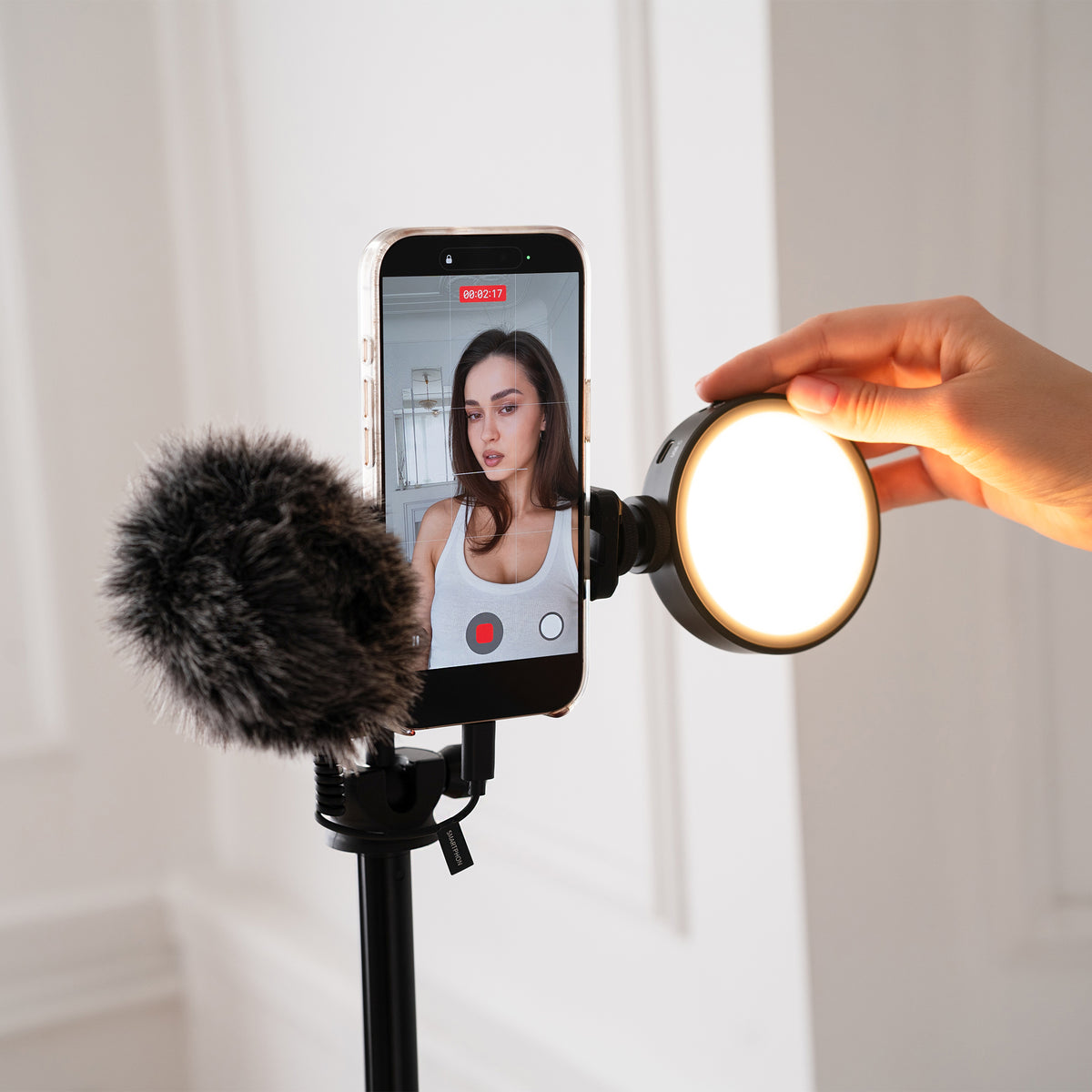 A smartphone video call setup with a phone on a stand, a furry microphone windscreen, and a hand holding a round ring light next to it. The phone screen shows a woman's video feed.