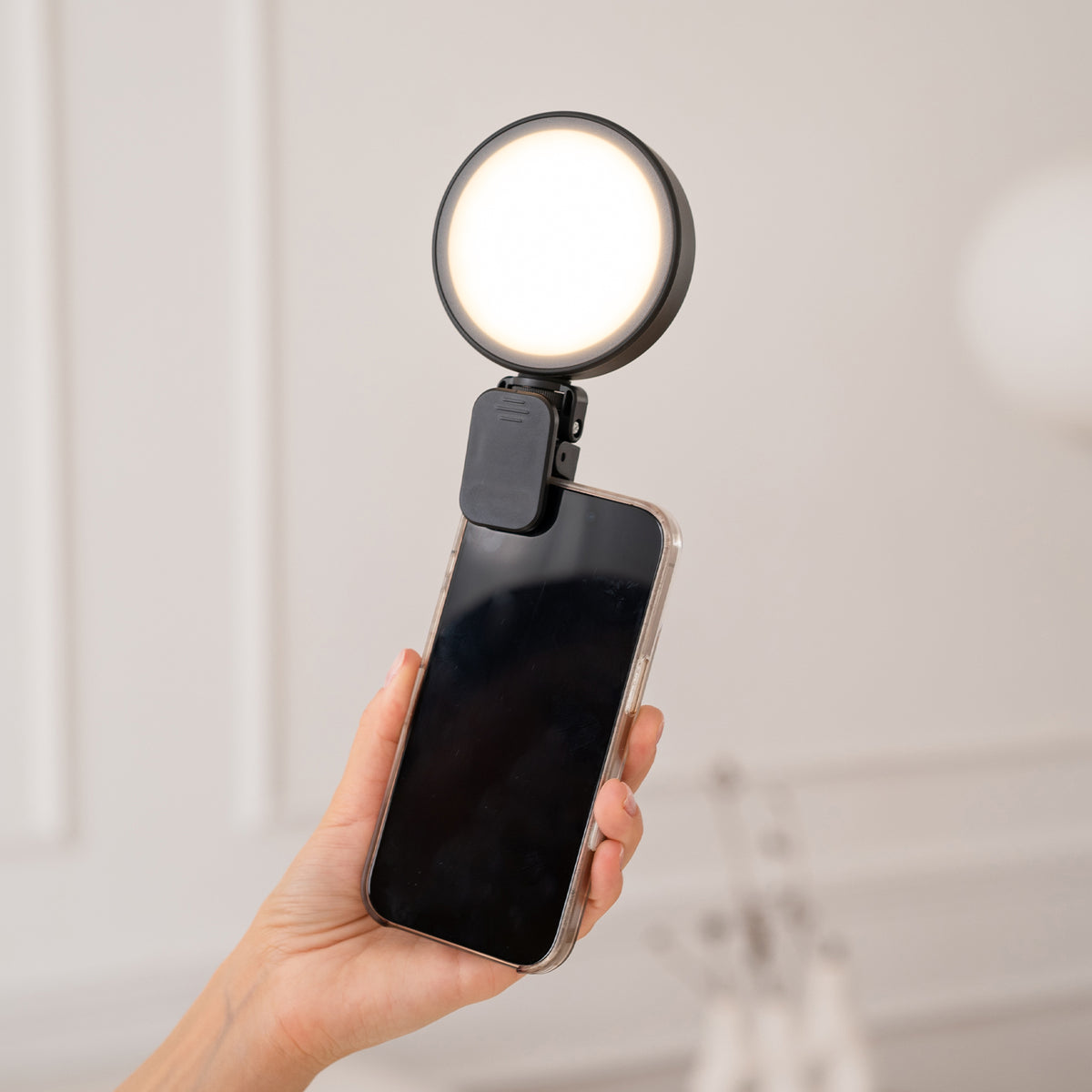 A hand holding a smartphone with a black clip-on ring light attached to the back, shown against a softly blurred indoor background.