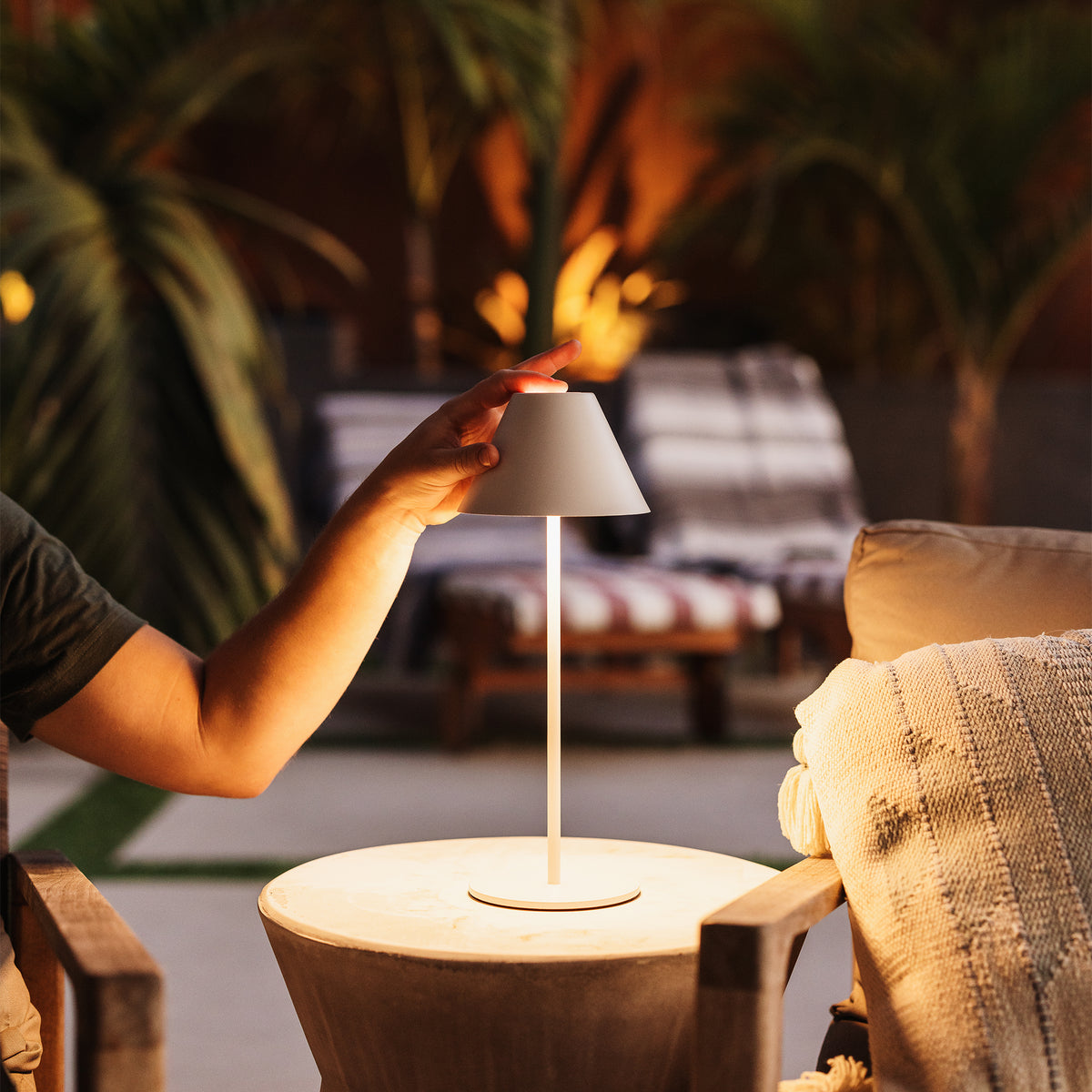 A modern table lamp with a white conical shade and slim white stem sits on a round side table in a warm, dimly lit patio setting. A person's hand is reaching toward the lamp, with lounge chairs and palm trees blurred in the background.