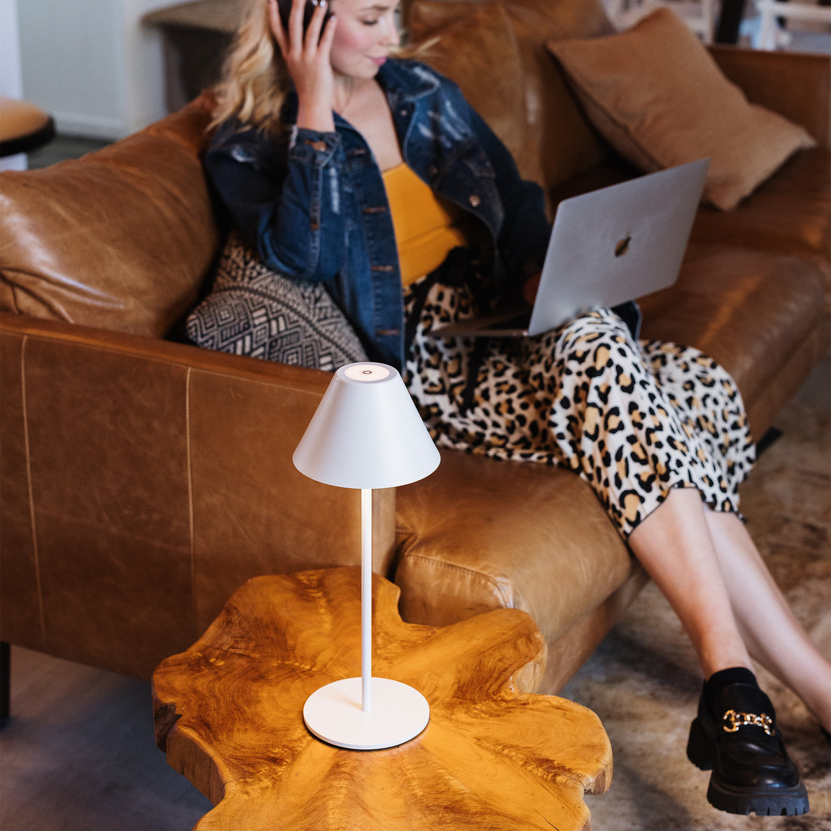 A white table lamp with a conical shade and slim stem sits on a rustic wood slab side table in a living room. A woman in a denim jacket, yellow top, and leopard-print skirt sits on a brown leather couch using a laptop.
