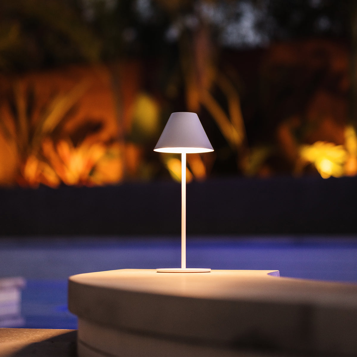 A modern minimalist table lamp with a white conical shade and slim white stem, glowing with warm light, set on a stone pedestal in front of a blurred outdoor poolside background at night.