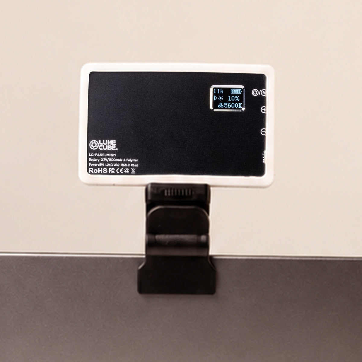 A Lume Labs light meter device with a white frame and black faceplate, clipped onto a dark surface against a light beige wall.