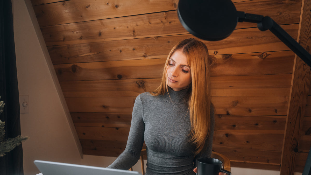 A woman with long strawberry-blonde hair wearing a gray turtleneck sweater sits in front of a wooden slat wall, looking down at a laptop screen. A black studio microphone on an adjustable arm is visible in the upper right, and she holds a black mug.